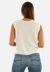 Maglioncino senza maniche beige chiaro con una finitura morbida e testurizzata. Orlo a coste e scollatura bassa alle ascelle. Indossato con jeans blu dotati di tasche posteriori.