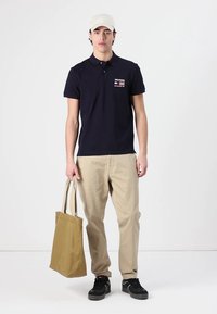 Marineblå polo skjorte med korte ærmer og merkemerker, beige bukser, sorte sneakers, og bærer en lysebrun håndveske, iført en lys caps.