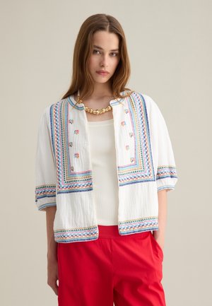 White embroidered jacket with colorful geometric patterns and blue trim. Worn over a fitted top, paired with red pants. Short sleeves, open front.