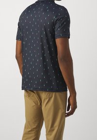 Man wearing navy blue polo shirt with small green golfer motifs and tan pants, shown from behind against white background.