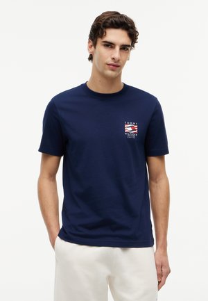 Νεαρός άνδρας που φοράει μπλε ναυτικό T-shirt Tommy Hilfiger με λογότυπο στο στήθος, συνδυασμένο με λευκό παντελόνι, στέκεται μπροστά σε απλό φόντο.