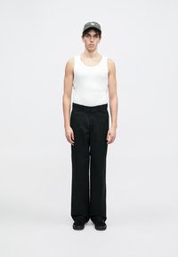 Sorte vide bukser kombinert med en hvit singlet. Modellen har på seg en grå cap og svarte joggesko. Minimalistisk design mot en ensfarget bakgrunn.