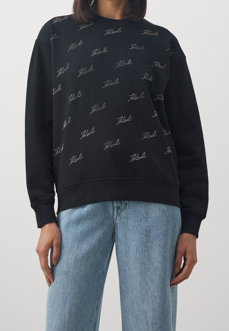 Personne portant un sweat-shirt noir avec un motif de texte argenté "Zad" et un jean en denim bleu clair, debout devant un fond clair uni.
