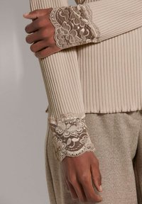 Beige geripptes Langarmoberteil mit aufwendigen Spitzenärmeln, die florale Muster aufweisen. Die glatte Textur des Ärmels kontrastiert mit dem Spitzen-detail.