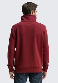 Bordeauxkleurige pullover sweatshirt met een hoge kraag, ribgebreide manchetten en zoom, en een gladde textuur. Bevat een logo patch op de linkermouw.