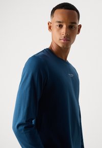 Jonge man met kort haar, gekleed in een donkerblauw, langemmouw shirt met een klein wit logo op de borst, staande tegen een effen lichte achtergrond.