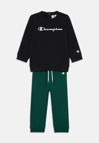 Svart hettegenser med hvit "Champion"-logo og grønne joggebukser med snøring og rødt logoakcent på venstre ben.
