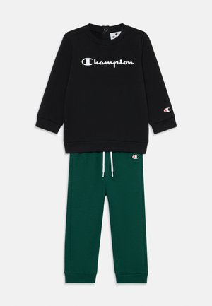 Czarny sweter z białym logo "Champion" i zielone spodnie dresowe ze sznurkiem oraz akcentem w kolorze czerwonym na lewym nogawce.