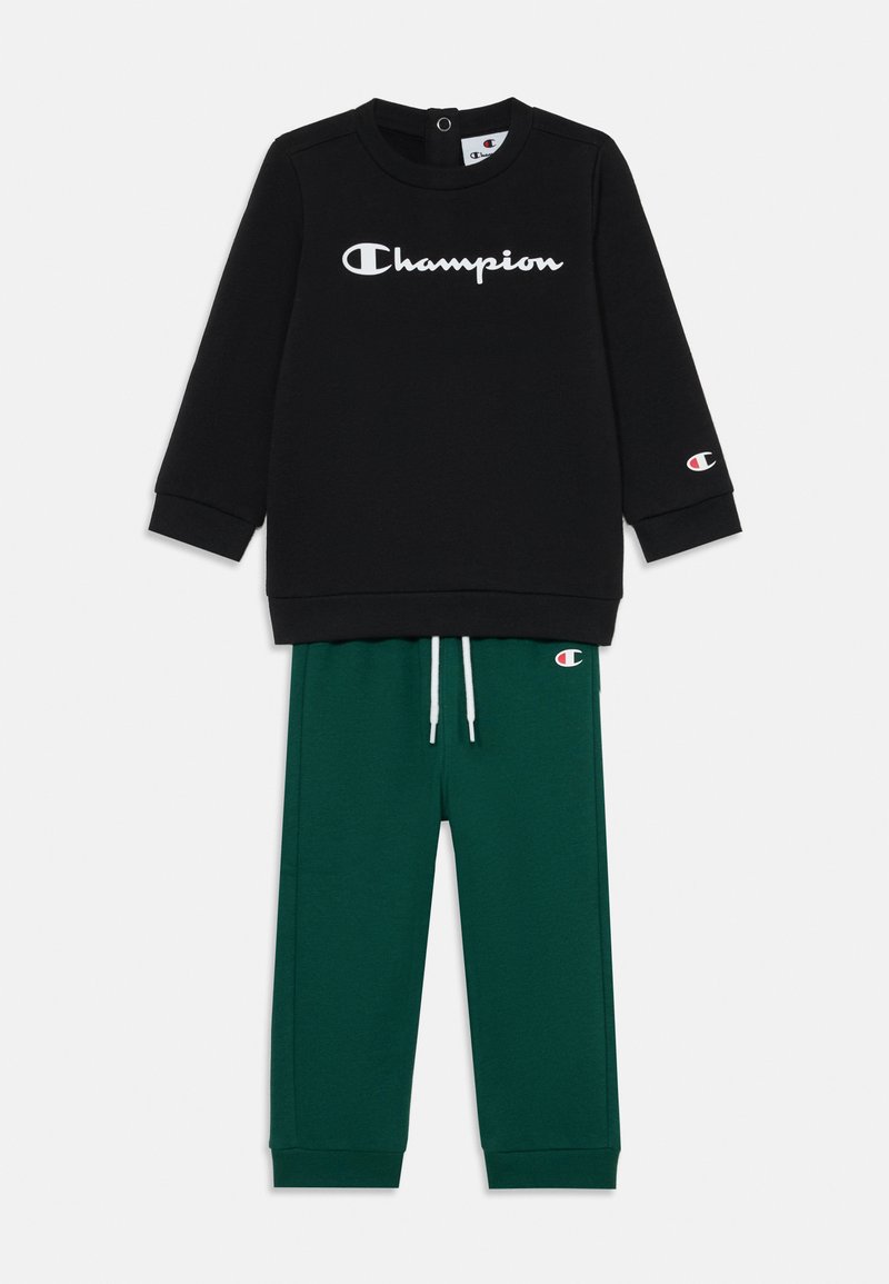Svart hettegenser med hvit "Champion"-logo og grønne joggebukser med snøring og rødt logoakcent på venstre ben.