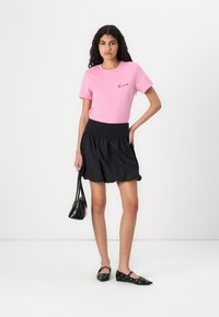 T-shirt en coton rose avec logo en script noir, associé à une jupe bouffante noire avec une taille élastique et des ballerines noires assorties.