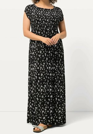 Kobieta w czarnej sukience maxi z białymi abstrakcyjnymi plamami, z krótkimi rękawami, w beżowych sandałach wsuwanych, stoi z złączonymi dłońmi.