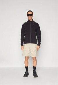 Mustast fliisi jakk, beežid lühikesed püksid, mustad sokid ja tennised. Jakil on ees tõmblukk ja logo detail. Tumedad päikeseprillid lõpetavad välimuse.