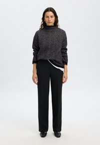 Črno-beli pulover z geometrijskim cik-cak vzorcem, z visokim ovratnikom, kombiniran s črnimi hlačami po meri in črnimi čevlji brez zavezovanja. Enostavna, teksturirana zasnova.