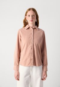 Light pink long-sleeve shirt with a collared neckline, button-down front, and subtle ribbed texture. Paired with white trousers.