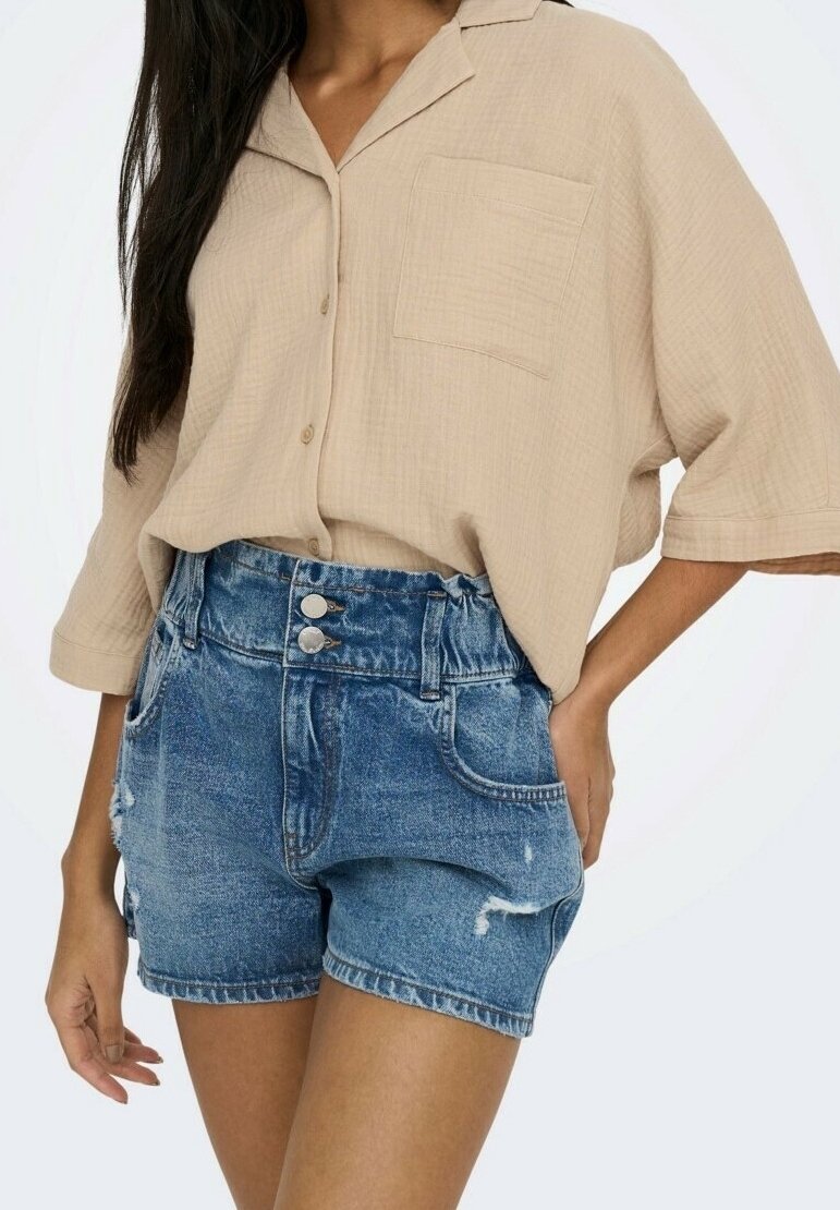 Chemise beige à manches courtes avec un bouton et une poche, associée à un short en denim bleu à taille haute, présentant des détails déchirés et une fermeture à double bouton.