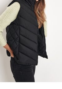 Černá prošívaná vesta s diamantovým vzorem, vysokým límcem a zipovým uzávěrem. V kombinaci se světle zeleným odstínem dlouhým rukávem s ribovaným vzorem.