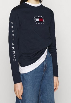 Osoba ubrana w granatową koszulę Tommy Jeans z długim rękawem, z logo na piersi i rękawach, w połączeniu z niebieskimi dżinsami i białą podkoszulką.