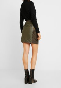 Jupe mini en cuir vert olive avec un panneau latéral en imprimé serpent texturé, présentant des détails en zip argenté, associée à des bottines noires.