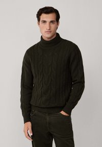 Man som bär en mörkgrön kabelstickad polotröja och matchande mörkgröna byxor, stående med ena handen i fickan.