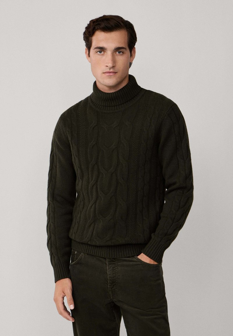 Man som bär en mörkgrön kabelstickad polotröja och matchande mörkgröna byxor, stående med ena handen i fickan.