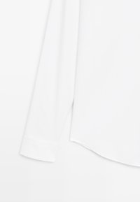 Camicia bianca a maniche lunghe con una texture liscia, polsini abbottonati e orlo curvo. Il materiale sembra leggero e traspirante.