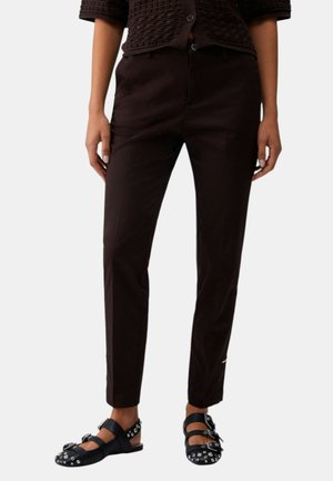 Donna che indossa pantaloni neri aderenti, top nero strutturato a maniche corte e sandali neri con borchie e fibbie argentate, in piedi su sfondo bianco.