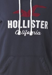 Marinblå huvtröja med vita snören. Har en röd broderad fågel-logotyp och texten "HOLLISTER California" i vitt. Inkluderar en framficka.