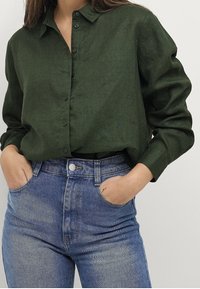 Camicia in lino verde scuro con bottini frontali, colletto a punta e maniche arrotolate, abbinata a jeans azzurri a vita alta.