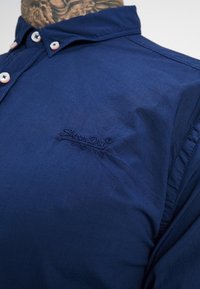 Námořnická modrá košile s knoflíky vyrobená z měkké látky, se světlými knoflíky a vyšitým logem na levé hrudi.