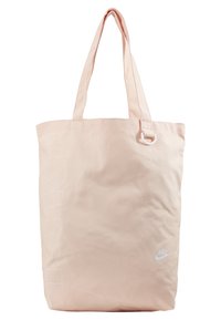 Jasnoróżowa torba tote wykonana z materiału, z dwiema długimi rączkami, białym logo Nike i pionowym wzorem tekstowym po jednej stronie.