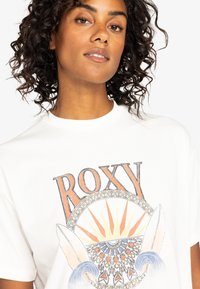 Biała bawełniana koszulka z kolorowym wzorem graficznym przedstawiającym słowo "ROXY", deski surfingowe, promienie słońca oraz motywy fal w odcieniach pomarańczowym i niebieskim.
