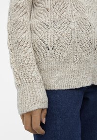 Pull beige en tricot avec un motif chevron texturé sur la manche et le corps, porté avec un jean en denim bleu foncé.