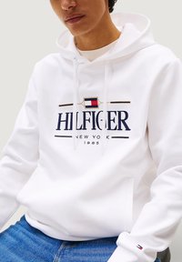 Felpa bianca con logo frontale "HILFIGER NEW YORK", caratterizzata da dettagli in blu navy e rosso. Cappuccio con cordini, tasca a marsupio e polsini a coste.
