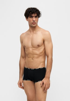 Ung mand med krøllet mørkt hår iført sorte boxershorts med hvid logo-linning på ensartet lys baggrund.