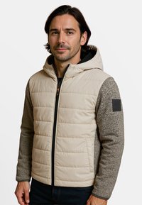 Lichtbeige pufferjack met een grijze gebreide capuchon en mouwen. Het heeft een ritssluiting aan de voorkant, zijzakken en een logo patch op de mouw.