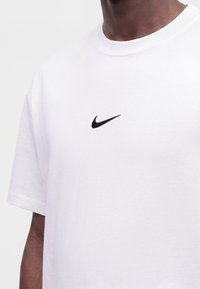 Άτομο φοράει ένα απλό λευκό t-shirt με ένα μικρό μαύρο λογότυπο Nike Swoosh κεντημένο στο στήθος.