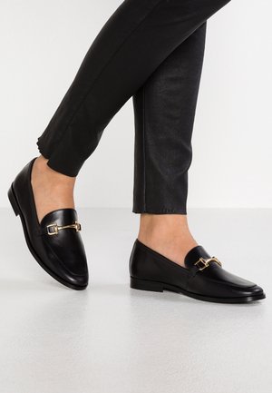 Personne portant des mocassins en cuir noir avec des boucles dorées et un pantalon noir ajusté, debout sur un sol clair et lisse.