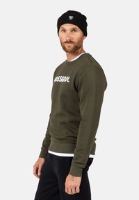 Felpa verde oliva con testo bianco "ROSSIGNOL", polsini a costine e scollatura rotonda. Indossata con un berretto nero e pantaloni neri.