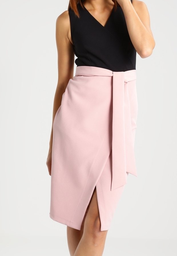 Femme portant un top noir sans manches associé à une jupe portefeuille rose pâle longueur genou avec une large ceinture nouée à la taille.