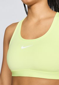 Ανοιχτό πράσινο αθλητικό σουτιέν κατασκευασμένο από απαλό υλικό με στρογγυλή λαιμόκοψη και λευκό λογότυπο Nike στο κέντρο. Απλός σχεδιασμός χωρίς επιπλέον μοτίβα.
