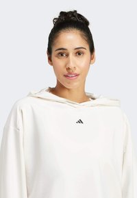 Weißer Kapuzenpullover aus weichem Stoff mit einer Kordelzugkapuze und einem kleinen schwarzen adidas-Logo auf der Brust. Glatte Textur und lässige Passform.