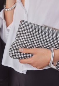 Hand houdt een zwarte clutch bedekt met strass-steentjes vast, draagt een wit overhemd, een zilveren armband en aan de andere hand een diamantbezette ring.