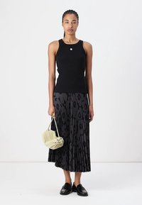 Top negru fără mâneci asortat cu o fustă plisată neagră, cu un model subtil. Modelul ține o geantă verde deschis și poartă loafers negri.