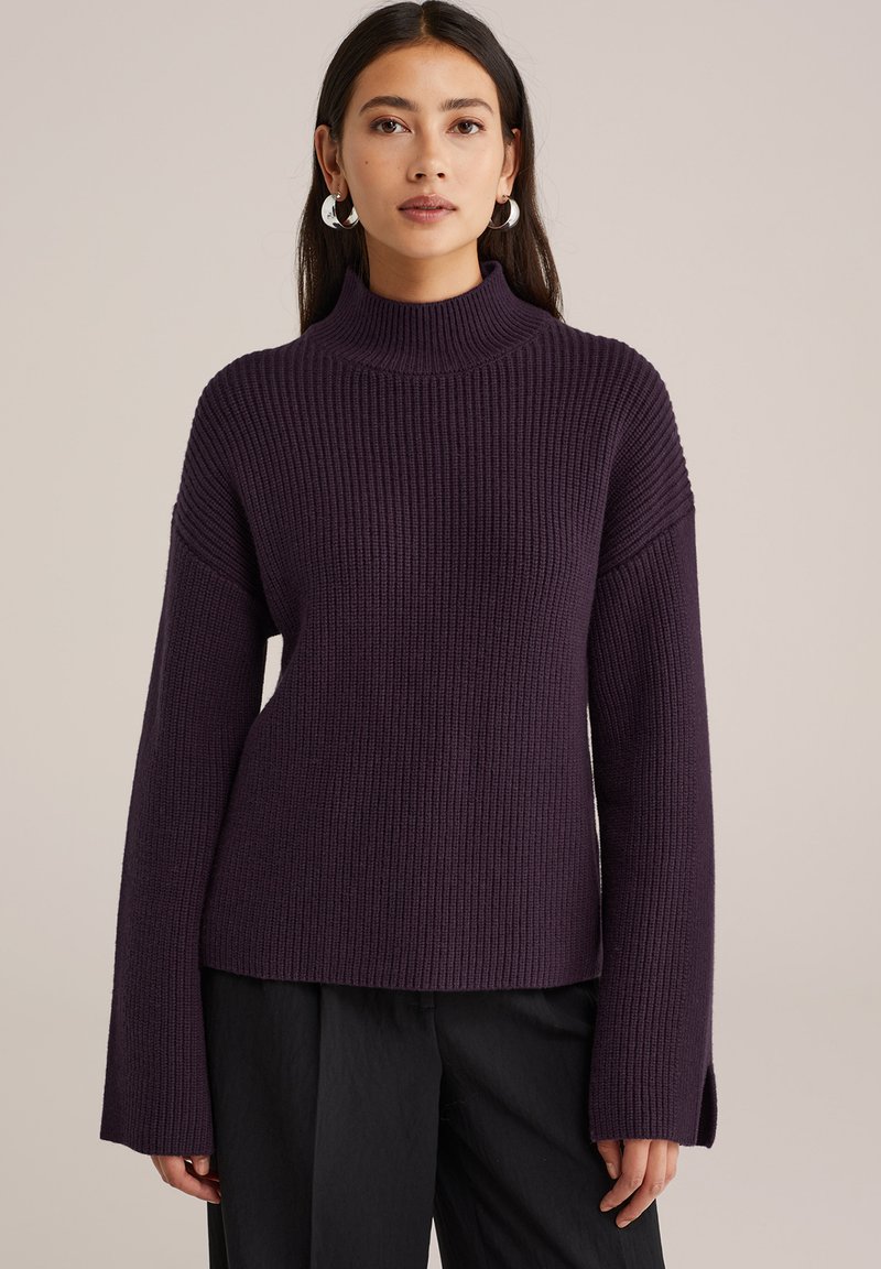 Femme portant un pull torsadé, violet foncé à col roulé avec des manches larges et un pantalon noir, se tenant devant un fond beige uni.
