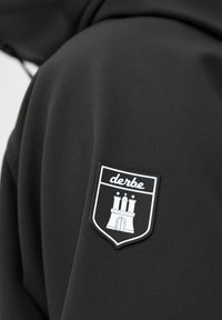 Zwarte hoodie met een gestructureerde stof en een opvallende zwarte schildpatch met een kasteelontwerp en de merknaam "derbe" in het wit.