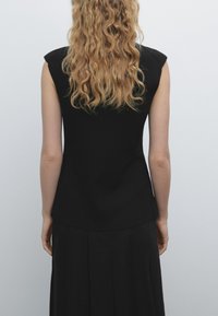 Black sleeveless top with structured shoulder design, smooth texture, and a fitted silhouette, paired with a pleated black skirt.