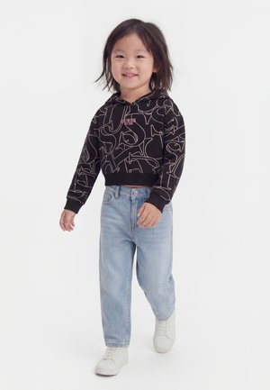 Lille barn går, iført sort mønstret hættetrøje, lyseblå jeans og hvide sneakers mod en ensfarvet hvid baggrund.