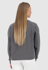 Felpa grigia con scollo rotondo, maniche raglan, polsini a coste e una texture morbida, indossata con pantaloni bianchi, vista da dietro.