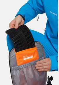 Ryggbeskyttelse i svart med teksturert overflate, lagret i et grått og oransje rom. En person som har på seg en blå jakke holder den.