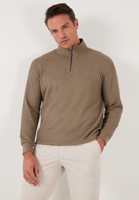 Pull en maille côtelée marron avec fermeture éclair, à manches longues et col montant, présentant une texture subtile et un accent de zip noir. Associé à un pantalon clair.